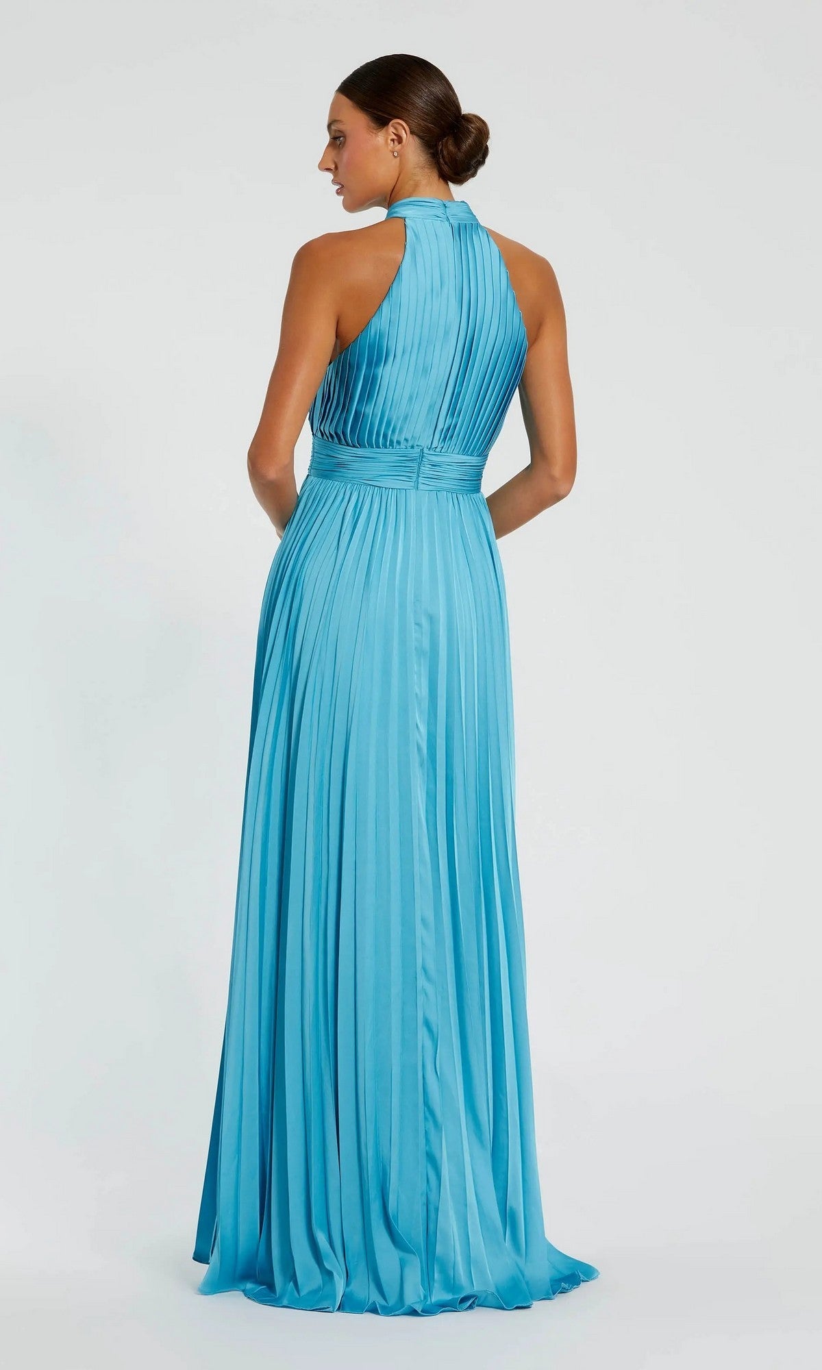 A woman stands facing away against a plain white background, her hair styled in a low bun, wearing the Mac Duggal 26992 long pleated A-line formal dress in light blue with a high neckline.